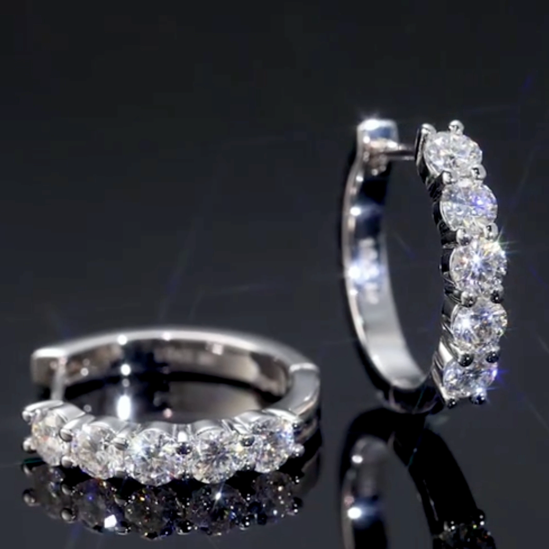 Pair of silver hoop earrings with clear gemstones on a reflective black surface.